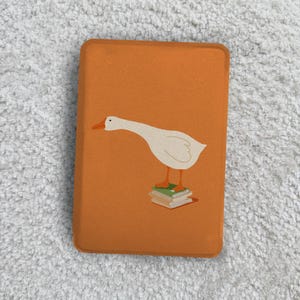 May include: A rectangular orange e-reader case featuring a graphic illustration of a white goose with an orange beak and feet standing on a small stack of three books. The case has rounded corners and a smooth, solid orange background color.