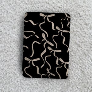 May include: A rectangular object with a black background featuring a repeating pattern of light beige ribbon bows. Each bow has long, flowing, curved tails that extend across the surface, creating a continuous and interconnected design.