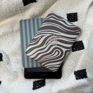 Puede incluir: Un lector de libros electrónicos Kindle negro con una funda a rayas azules y grises y una funda con estampado de cebra marrón y blanco. La funda con estampado de cebra tiene el texto "for girls read". Los artículos están sobre una superficie con estampado blanco y negro.