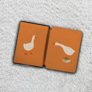 May include: An open book with an orange cover featuring illustrations of white geese. One goose is walking, and the other is standing on a stack of books. The book has a black border.