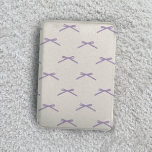 以下が含まれることがあります： オフホワイトの長方形の携帯電話ケース。ラベンダー色のリボンの繰り返し模様があります。リボンは表面全体に均等に配置されています。ケースは角が丸く、滑らかな仕上がりです。