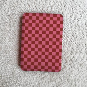 Peut inclure: Un motif à carreaux rose et marron sur un objet rectangulaire. Le motif est une grille de carrés dans deux nuances de rose et de marron. L'objet est posé sur un fond blanc texturé.