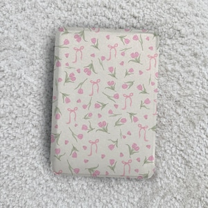 May include: A rectangular object featuring a light beige background with a repeating pattern of small pink tulips with green stems and leaves, interspersed with delicate pink ribbon bows scattered across the entire surface.