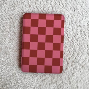 Peut inclure: Un étui rectangulaire avec un motif à carreaux rose et marron. L'étui a des coins arrondis et est posé sur un fond blanc texturé. L'étui est probablement pour un téléphone ou une tablette.