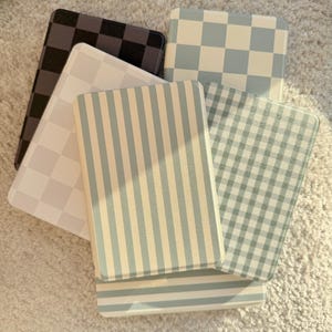May include: A selection of rectangular coasters with diverse patterns. Designs include checkered, striped, and gingham patterns in shades of black, white, and blue. The coasters are arranged on a light-coloured, textured surface.