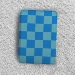 Peut inclure: Un objet rectangulaire avec un motif à carreaux bleu et turquoise. Le motif présente une grille de carrés alternés en deux nuances de bleu. Il a des coins arrondis et est posé sur un fond blanc texturé.