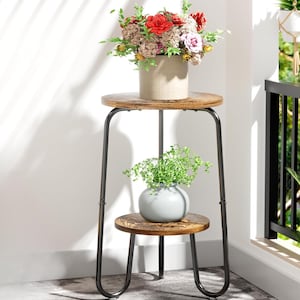 May include: A two-tiered plant stand with a distressed wood finish and black metal frame. The top tier holds a beige pot with colorful flowers, while the bottom tier displays a gray vase with greenery. The stand is approximately 24 inches tall.