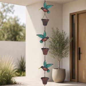 May include: A decorative rain chain featuring three hummingbirds with teal wings and brown bodies, each suspended above a small, woven-style basket. The chain is designed for outdoor use and adds a touch of nature-inspired charm to any space.