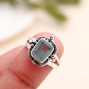May include: A silver ring featuring a rectangular, light blue gemstone. The ring has an ornate design with decorative elements around the stone. The band splits into two at the top, adding to its unique style. A piece of jewelry.