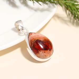 May include: A teardrop-shaped pendant with a polished, reddish-brown gemstone set in a silver bezel. The gemstone has a mix of colors, including red, orange, and cream. The pendant hangs from a silver bail.