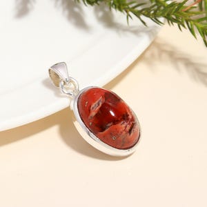 May include: Oval pendant featuring a red and brown stone, set within a silver-coloured metal frame. The pendant includes a loop at the top for a chain. The stone displays a marbled pattern.