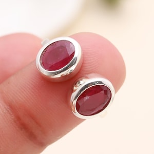 May include: A pair of silver stud earrings, each featuring a vibrant oval-cut ruby gemstone. The rubies are a deep red color, set in a polished silver bezel. The earrings are displayed against a soft, neutral background.
