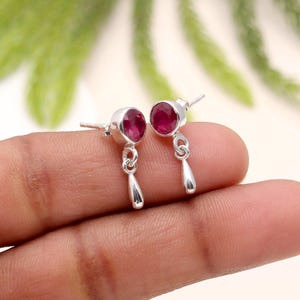 Puede incluir: Un par de pendientes de plata con una gema redonda de color rojo intenso y un dije colgante en forma de lágrima. Los pendientes se muestran sobre un fondo borroso, mostrando el diseño y el color de la joya.