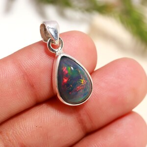 May include: A teardrop-shaped pendant with a black opal gemstone. The opal displays vibrant flashes of red, green, and yellow within its dark surface. The pendant is set in a silver metal frame with a loop for a chain.