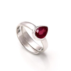 May include: A silver ring featuring a pear-shaped, deep red gemstone. The ring has a smooth, polished band that curves around the finger, with the gemstone set in a bezel setting. The ring is displayed against a white background.