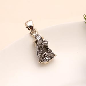 May include: A silver pendant featuring a rough, textured, dark gray meteorite. The pendant has a silver bail for a chain and small silver accents at the base. The meteorite has a unique, irregular shape.