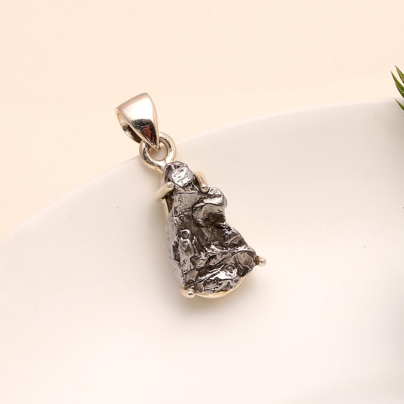 May include: A silver pendant featuring a rough, textured, dark gray meteorite. The pendant has a silver bail for a chain and small silver accents at the base. The meteorite has a unique, irregular shape.