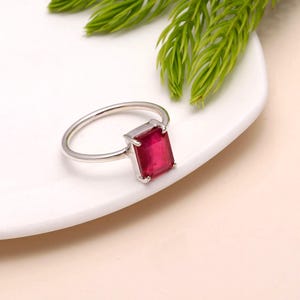May include: A silver ring with a rectangular, deep red gemstone. The ring is set on a white surface with green foliage in the background. The gemstone is the focal point, showcasing its vibrant color and shape.