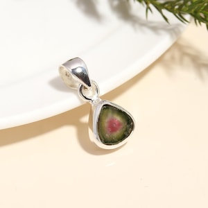 Puede incluir: Colgante de plata con una piedra preciosa de turmalina sandía. La piedra preciosa tiene forma triangular con una capa exterior verde y un centro rosa. El colgante está sobre un plato blanco.