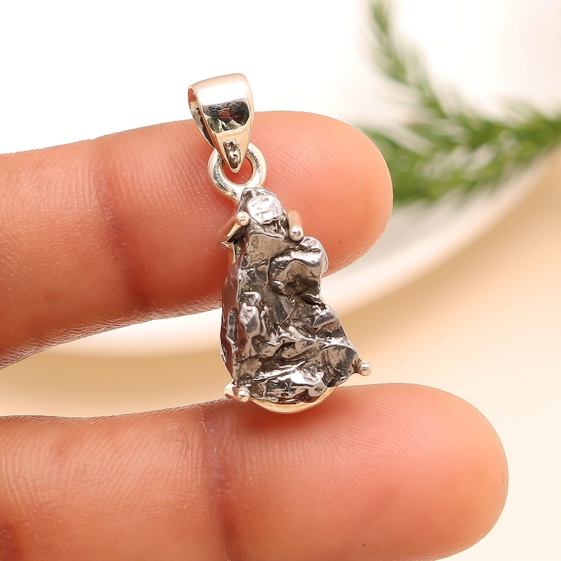 May include: A silver pendant with a unique, textured, irregular shape. The pendant is attached to a silver bail. The overall design has a rugged, natural aesthetic, suggesting a gemstone or mineral. The pendant is held between two fingers.