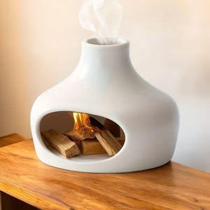 May include: A white ceramic fireplace with a rounded body and an oval opening. Inside, burning wood emits flames and smoke. The fireplace is placed on a wooden surface.