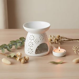 May include: A white ceramic essential oil diffuser with a floral cut-out design. A small, white candle holder with a lit tea light is next to the diffuser. Dried flowers and greenery are arranged around the objects on a light wood surface.