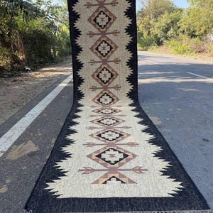 Puede incluir: Una alfombra de pasillo rectangular larga con un centro color crema que presenta un patrón repetitivo de formas geométricas de diamante y motivos de flechas en negro, tostado y marrón, bordeada por un borde negro dentado y flecos cortos.