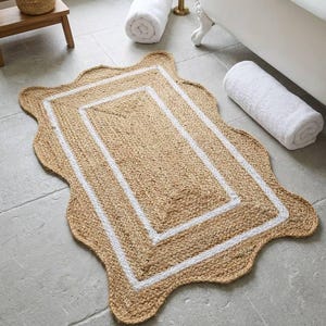 May include: A rectangular bath mat with a scalloped edge, made of natural jute fiber. The mat features a white border and a central rectangular design. Rolled white towels and a wooden stool with bath accessories are in the background.