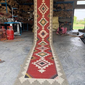 Puede incluir: Una alfombra larga y rectangular con un campo central rojo con patrones geométricos de diamantes en blanco, negro, amarillo y verde. La alfombra tiene un borde gris y flecos en los extremos. La alfombra se exhibe en un taller.