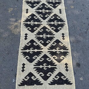 Puede incluir: Una alfombra de pasillo rectangular con un patrón geométrico de diamantes negros repetidos sobre un fondo color crema. La alfombra tiene una textura tejida y está rematada con flecos cortos de color gris oscuro en ambos extremos.