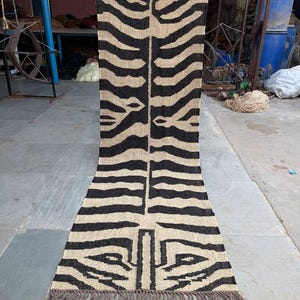 May include: A long, rectangular rug with a zebra stripe pattern in black and beige. The rug has a fringed edge and is held up vertically. The design is symmetrical, with the stripes mirroring each other down the center.