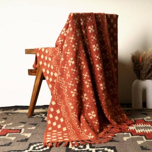 May include: A rust-colored textile throw blanket featuring a repeating pattern of cream-colored geometric shapes, including diamonds, zigzags, and floral-like motifs. The fabric has a textured weave and is finished with fringe along the bottom edge.