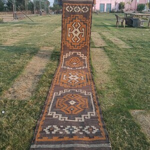 Puede incluir: Una alfombra larga y rectangular marrón con patrones geométricos en tonos naranja, blanco y gris. La alfombra se exhibe al aire libre sobre una zona de césped. El diseño presenta formas y bordes repetidos, con un borde con flecos.