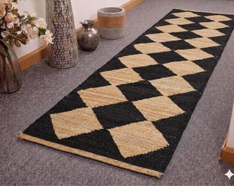 May include: A rectangular jute runner rug with a black and tan diamond pattern. The rug is placed on a gray surface, with decorative vases and a small pot in the background. The rug's design adds a touch of texture and visual interest to the space.