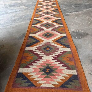May include: A long, rectangular runner rug with a geometric pattern in shades of orange, pink, green, and navy blue. The rug has a fringed edge and is likely made of woven material. The design features diamond and zig-zag shapes.