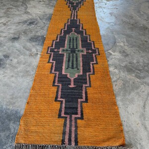 Puede incluir: Una alfombra larga y rectangular con un diseño geométrico. La alfombra presenta un fondo amarillo mostaza con un patrón central en negro, rosa y verde. La alfombra tiene flecos en ambos extremos.