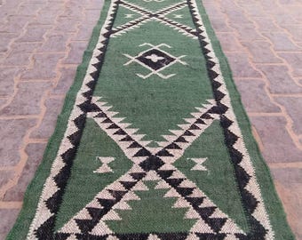 May include: Green and black geometric patterned runner rug with a rolled-up section at the top. The rug features a repeating diamond and triangle design in black and white against a green background. The rug has a fringe at the bottom.