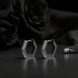 May include: A pair of matte grey hexagonal hoop earrings. The earrings are made of metal and have a hinged closure. The background is dark, with a blurred floral element and a textured surface.