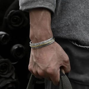 May include: Silver cuff bracelet featuring a feather design with gold-coloured detailing. The bracelet is worn on a wrist, highlighting the feather pattern. This piece of jewellery is a stylish accessory.