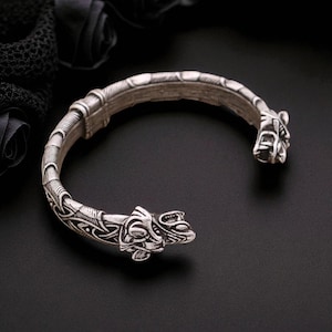 May include: Silver-toned cuff bracelet with intricate Viking-inspired designs. The bracelet features detailed carvings and ends with stylized animal heads. The open design allows for easy wear. The bracelet is displayed on a black surface.