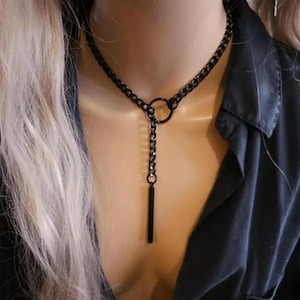 Puede incluir: Un collar de metal negro con un diseño de cadena. El collar presenta un colgante circular y un colgante de barra vertical. El collar lo lleva una persona con cabello rubio y una camisa negra.