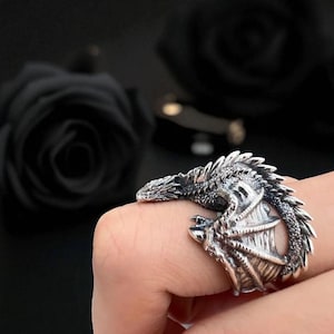 Könnte beinhalten: Ein silberner Drachenring mit aufwendigen Details. Der Drache umschlingt den Finger und weist Schuppen, Krallen und einen detaillierten Kopf auf. Der Ring ist vor einem dunklen Hintergrund platziert, der das metallische Finish hervorhebt.