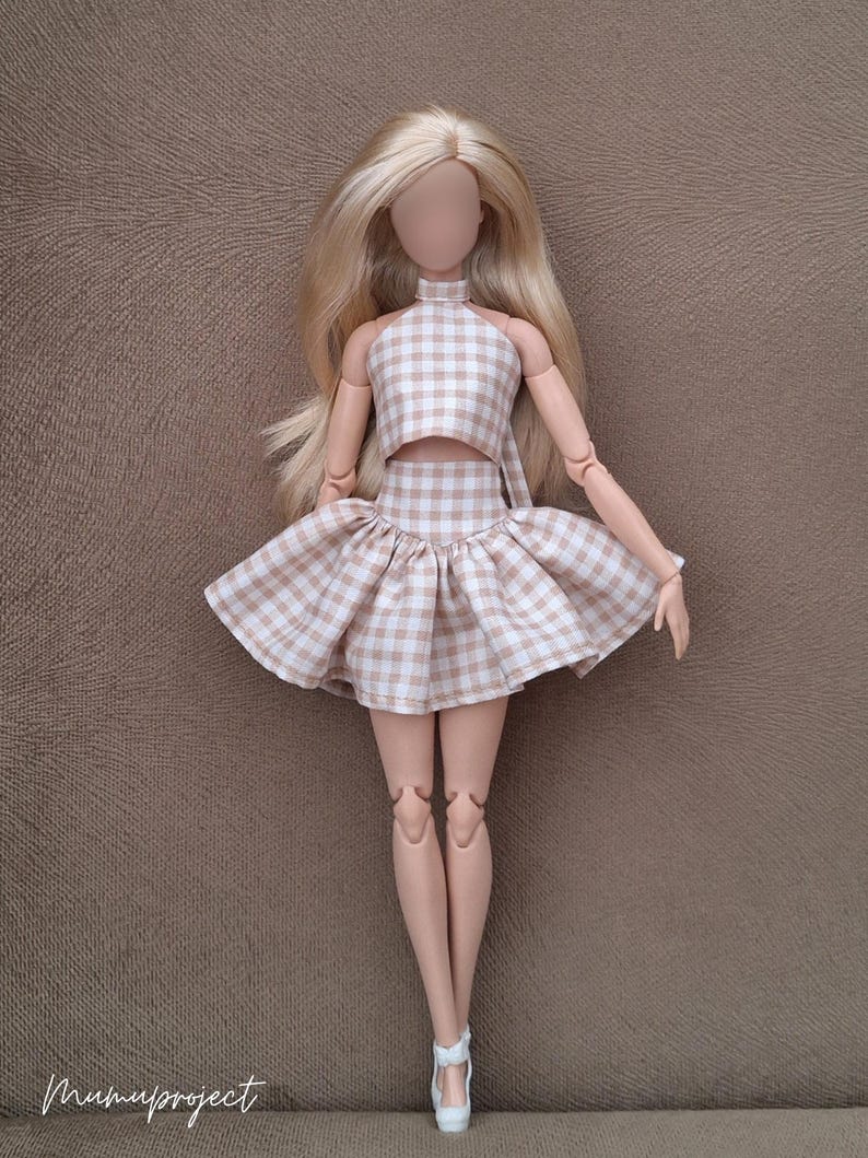 May include: A doll wearing a two-piece outfit with a halter top and a flared skirt. The outfit is a light brown and white gingham pattern. The doll has long blonde hair and white shoes. The doll is standing on a brown surface.