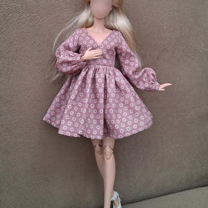 May include: A doll with long blonde hair wearing a mauve dress featuring a white daisy pattern, a V-neckline, and long puffed sleeves with cuffs. The doll has articulated joints and is wearing white shoes. The text mumuproject is visible.