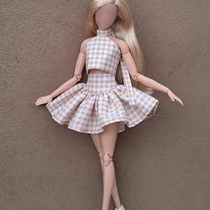May include: A doll wearing a two-piece outfit with a halter top and a flared skirt. The outfit is a light brown and white gingham pattern. The doll has long blonde hair and white high heels. The doll is standing on a brown surface.