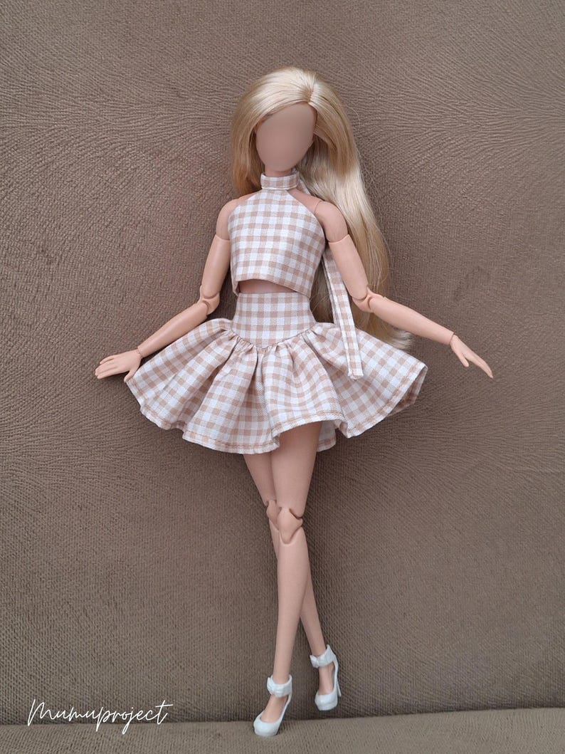 May include: A doll wearing a two-piece outfit with a halter top and a flared skirt. The outfit is a light brown and white gingham pattern. The doll has long blonde hair and white high heels. The doll is standing on a brown surface.