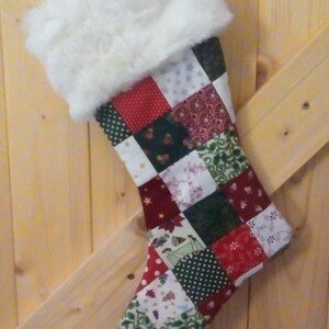 Op de afbeelding: Een handgemaakte kerstsok met een patchwork-ontwerp. De sok heeft een witte imitatiebontmanchet en een lichaam dat bestaat uit verschillende stoffen vierkanten in rode, groene en witte patronen. Het hangt tegen een houten achtergrond.