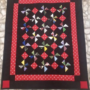 May include: A handmade quilt with a black background, red accents, and colorful pinwheel designs. The quilt features a red border with white floral patterns and red corner squares. The pinwheels are made of various patterned fabrics.