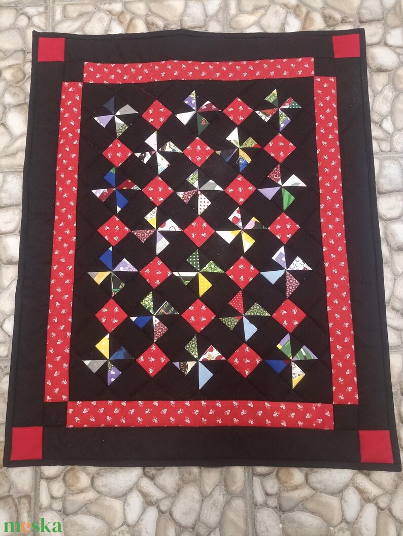 May include: A handmade quilt with a black background, red accents, and colourful pinwheel designs. The quilt features a red border with white floral patterns and red corner squares. The pinwheels are made of various patterned fabrics.