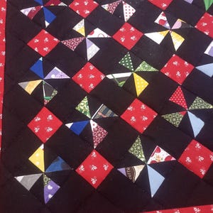 May include: Close-up of a handmade quilt with a black background. The quilt features a repeating pattern of red squares and colourful pinwheel designs. The border is red with a white floral pattern.